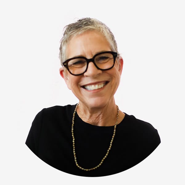 Smiling older woman with short gray hair, glasses, and a black top with a beaded necklace, photographed against a plain white background—ideal for Service Dog Certifications profiles.