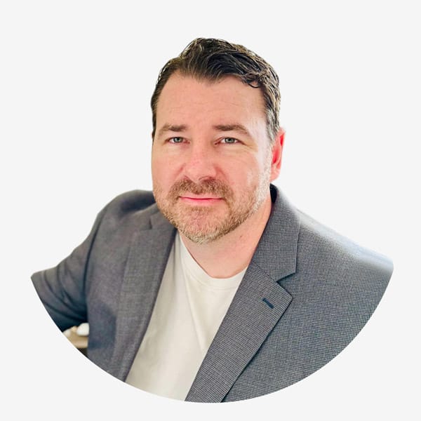 A man with short brown hair and a trimmed beard wearing a gray blazer over a white shirt stands against a plain white background, holding documents related to Service Dog Certifications.