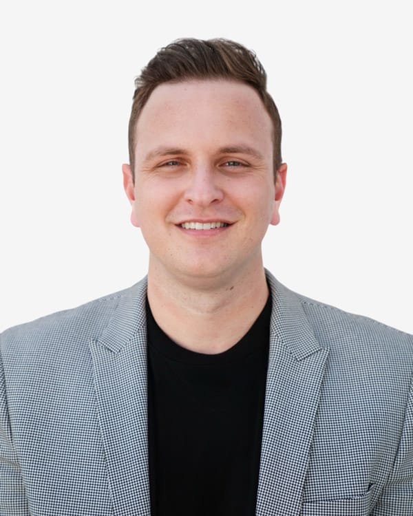 A man with short brown hair, wearing a light gray checked blazer and a black shirt, smiles at the camera against a plain white background.