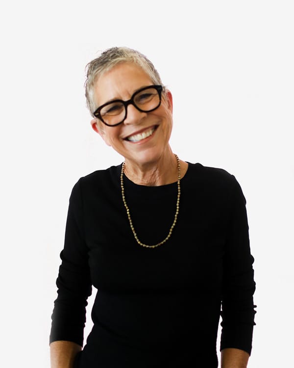 Smiling older woman with short gray hair, glasses, and a black long-sleeve top wearing a beaded necklace against a plain white background.